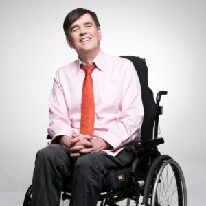 tim with a big smile sitting in wheelchair, and a pale business shirt and a red tie, fingers crossed. Adelaide Fringe Accessibility