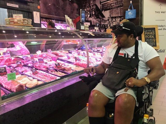 Ray is shown in his wheelchair. To his left is a long and high display case at a butcher's shop.