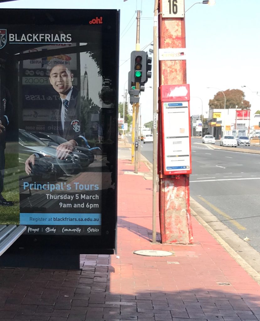 from the left to the right 1) a standard bus stop with advertising behind glass and a bench seat 2) background is traffic light showing green 3) a bus stop close to the road 4) the road. The bus stop hides people going in opposite directions