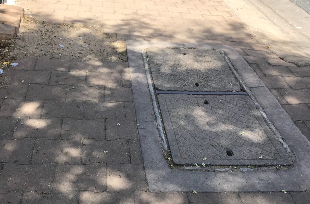 A pavement on a busy road/ 1/3 of the walkway is dirt and sand, the middle section is a utility hole with a cover, and the third part are pavers