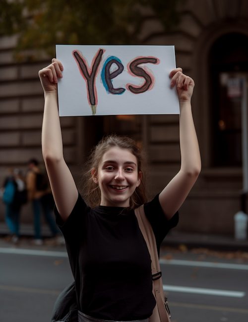 Vote YES in the Australian Voice Referendum - a young woman in a black shirt is holding a yes sign above her head