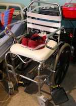 A lightweight metal shower chair with black wheels sits next to the bed. On the seat cushion rests a neatly placed pair of red running shoes, ready for use. sleeping after spinal cord injury