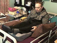 The man sits upright in bed, dressed in a gray jacket and black pants. His legs are supported by a thick gray vibrating cushion and brown pillows. He looks calm and comfortable, preparing for rest or a therapy routine. sleeping after spinal cord injury